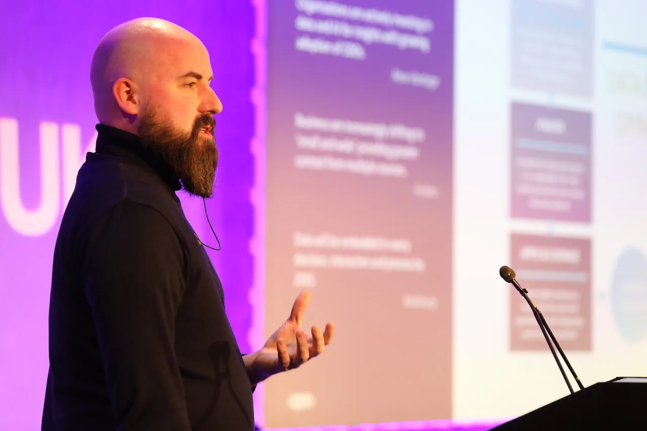 Christopher Howard presenting at a conference podium with presentation slides visible behind him